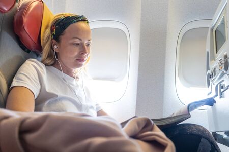 Casual young woman reading magazine and listening to music on airplane. Female traveler reading seated in passanger cabin. Sun shining trough airplane window.の写真素材