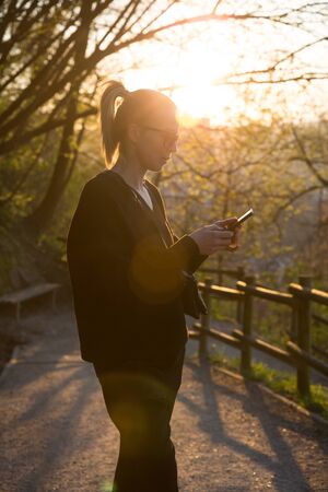 Backlit rear view of young woman talking on cell phone outdoors in park at sunset. Girl holding...