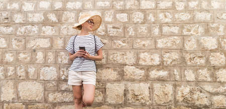 Beautiful young female tourist woman standing in front of old textured stone wall at old Mediterranean town, smiling, holding, smart phone to network on vacationes. Copy space.の写真素材