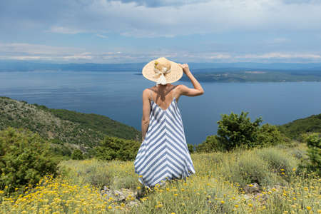 Rear view of young woman wearing striped summer dress and straw hat standing in super bloom of...