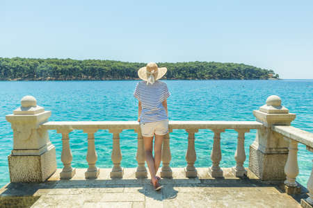 Rear view of woman wearing straw summer hat ,leaning against elegant old stone fence of coastal villa, relaxing while looking at blue Adriatic sea, on Losinj island Croatia.の写真素材