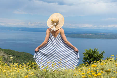 Rear view of young woman wearing striped summer dress and straw hat standing in super bloom of wildflowers, relaxing while enjoing beautiful view of Adriatic sea nature, Croatia.の写真素材