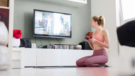 Attractive sporty woman working out at home, doing pilates exercise in front of television in small studio appartment. Social distancing. Stay healthy and stay at home during corona virus pandemic.の写真素材