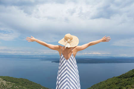 Rear view of young woman wearing striped summer dress and straw hat standing in super bloom of wildflowers, relaxing with hands up to the sky, enjoing beautiful view of Adriatic sea nature, Croatia.の写真素材
