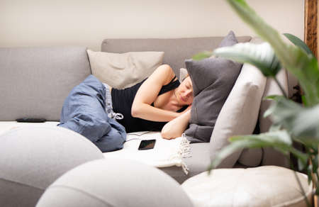 Tired young woman taking a nap at home lying on a comfortable sofa with big cushions in living room with a mobile phone lying next to her.の写真素材