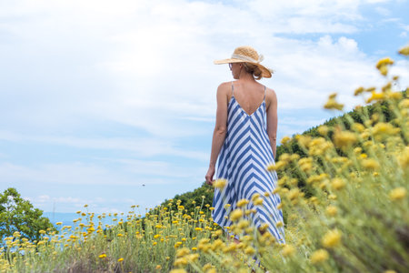 Young woman wearing striped summer dress and straw hat standing in super bloom of wildflowers, relaxing while enjoing beautiful nature of of Adriatic sea coastal nature of Croatia.の写真素材