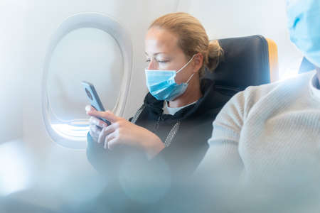 A young woman wearing face mask, using smart phone while traveling on airplane. New normal travel...