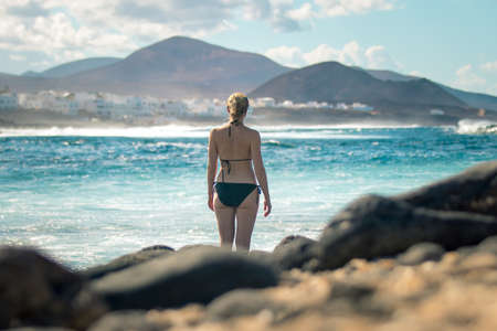 Female tourist at wild rocky beach and coastline of surf spot La Santa Lanzarote, Canary Islands, Spain. La Santa village and volcano mountain in background.の写真素材