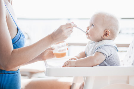 Mother spoon feeding her baby boy child in baby chair with fruit puree on a porch on summer vacations. Baby solid food introduction concept.の写真素材