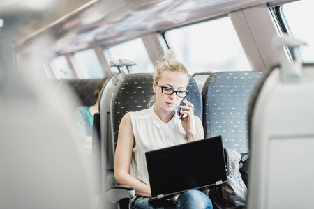 Businesswoman talking on cellphone and working on laptop while traveling by train. Business travel concept.の写真素材