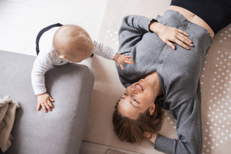 Happy family moments. Mother lying comfortably on childrens mat playing with her baby boy watching and suppervising his first steps. Positive human emotions, feelings, joyの写真素材