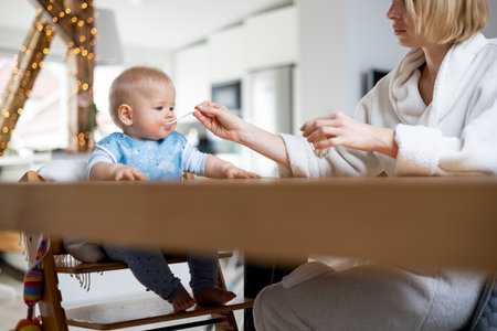 Mother wearing cosy bathrope spoon feeding her baby boy child in baby high chair with fruit puree at dinning table at home. Baby solid food introduction concept.の写真素材