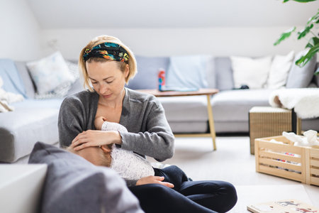 Young woman breastfeeding her infant baby boy casualy sitting on childs playing mat on living room...