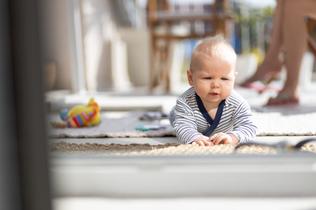Cute little infant baby boy playing with toys outdoors at the patio in summer being supervised by her mother seen in the background. Selective focus.の写真素材