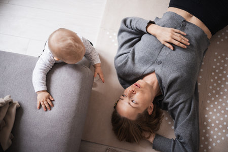 Happy family moments. Mother lying comfortably on childrens mat playing with her baby boy watching and suppervising his first steps. Positive human emotions, feelings, joyの写真素材