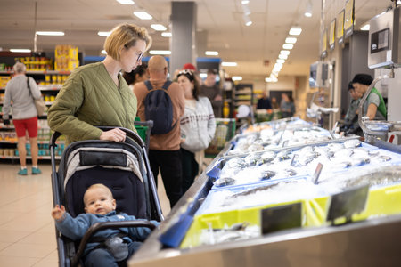 Casualy dressed mother choosing fish in the fish market department of supermarket grocery store with her infant baby boy child in strollerの写真素材