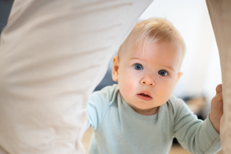 Portrait of adorable curious infant baby boy child taking first steps holding to fathers pants at...