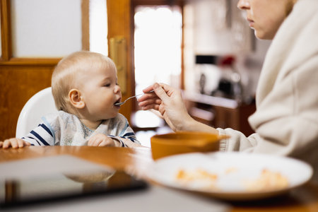 Mother spoon feeding her infant baby boy child sitting in high chair at the dining table in kitchen at home.の写真素材
