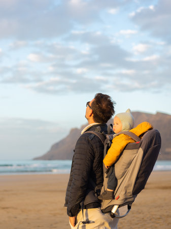 Father rising hands to the sky while enjoying pure nature carrying his infant baby boy son in backpack on windy sandy beach of Famara, Lanzarote island, Spain at sunset. Family travel conceptの写真素材