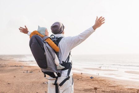 Young father rising hands to the sky while enjoying pure nature carrying his infant baby boy son in backpack on windy sandy beach. Family travel conceptの写真素材
