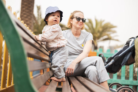 Young mother with her cute infant baby boy child on bench on urban childrens playground on warm summer dayの写真素材