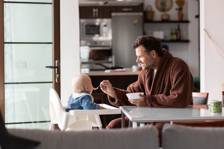 Father wearing bathrope spoon feeding hir infant baby boy child sitting in high chair at the dining table in kitchen at home in the morning.の写真素材