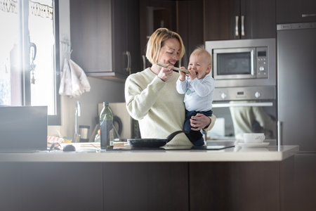 Happy mother and little infant baby boy cooking and tasting healthy dinner in domestic kitchen. Family, lifestyle, domestic life, food, healthy eating and people conceptの写真素材