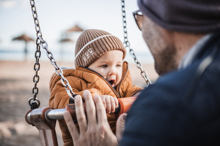 Father pushing hir cheerful infant baby boy child on a swing on sandy beach playground outdoors on nice sunny cold winter day in Malaga, Spainの写真素材