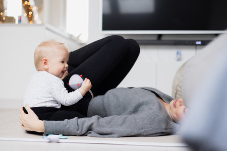 Happy family moments. Mother lying comfortably on childrens mat playing with her baby boy watching and suppervising his first steps. Positive human emotions, feelings, joyの写真素材