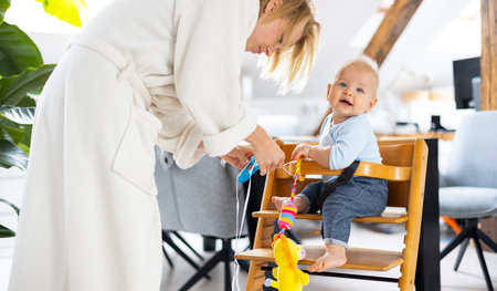 Happy infant sitting and playing with his toy in traditional scandinavian designer wooden high chair in modern bright atic home suppervised by his motherの写真素材