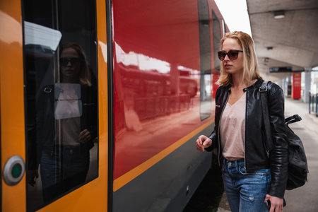 Young blond woman in jeans, shirt and leather jacket wearing bag and sunglass, embarking red modern speed train on train station platform. Travel and transportationの写真素材