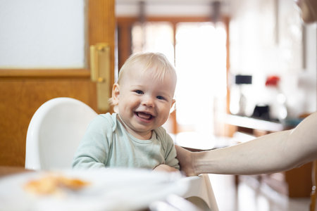 Adorable cheerful happy infant baby boy child smiling while sitting in high chair at the dining table in kitchen at home beeing spoon fed by his mother.の写真素材