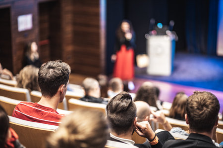 Business and entrepreneurship symposium. Female speaker giving a talk at business meeting. Audience in conference hall. Rear view of unrecognized participant in audience.の写真素材