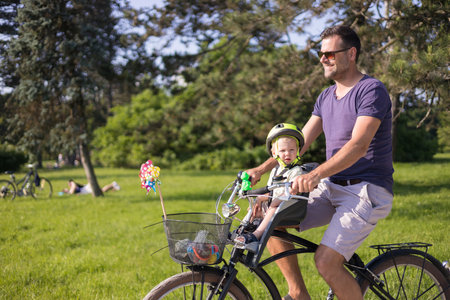 Look over there. Active family day in nature. Father and son ride bike through city park on unny summer day. A cute boy is sitting in front bicycle chair while father rides bicycle. Father son bondingの写真素材