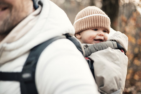 Sporty father carrying his infant son wearing winter jumpsuit and cap in backpack carrier hiking in autumn forestの写真素材