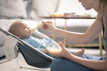 Mother spoon feeding her baby boy child in baby chair with fruit puree. Baby solid food introduction conceptの写真素材