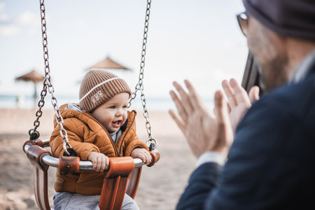 Father pushing hir cheerful infant baby boy child on a swing on sandy beach playground outdoors on nice sunny cold winter day in Malaga, Spainの写真素材