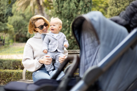 Mother sitting on bench in urban park, laughing cheerfully, holding her smiling infant baby boy child in her lap having baby stroller parked by their site.の写真素材