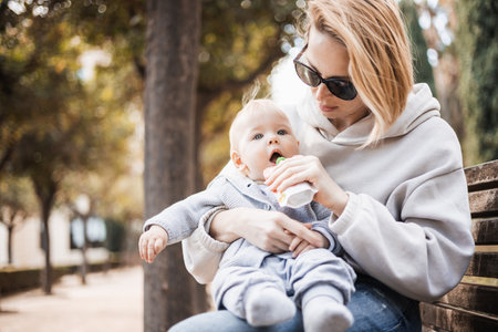 Mother sitting on bench in urban park, holding her infant baby boy child in her lap and feeding him baby food on nice spring sunny day.の写真素材