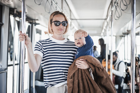 Mother carries her child while standing and holding on to bar holder on bus. Mom holding infant baby boy in her arms while riding in public transportation. Cute toddler traveling with motherの写真素材