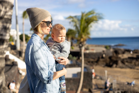 Fashinable mother holding her infant boy child in lap enjoing panoramic view along palm lined beach promenade of Puerto del Carmen, Lanzarote, Canary islands, Spainの写真素材