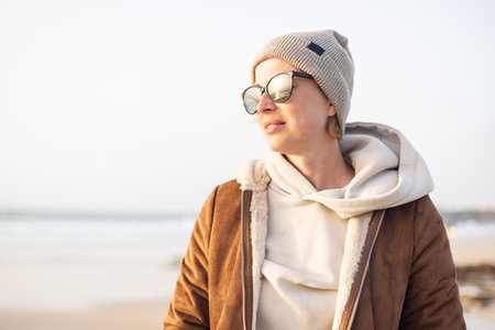 Portrait of young stylish woman wearing brown padded jacket, hoodie, wool cap and sunglasses on long sandy beach in springの写真素材