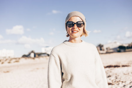 Portrait of young stylish woman wearing wool sweater, wool cap and sunglasses on long sandy beach in springの写真素材