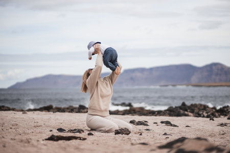 Mother enjoying winter vacations holding, playing and lifting his infant baby boy son high in the air on sandy beach on Lanzarote island, Spain. Family travel and vacations concept.の写真素材