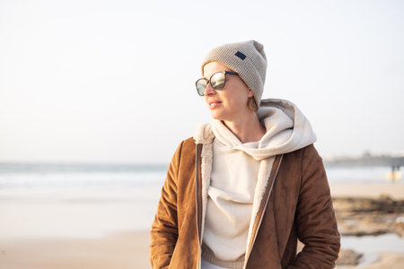 Portrait of young stylish woman wearing brown padded jacket, hoodie, wool cap and sunglasses on long sandy beach in springの写真素材