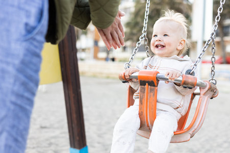 Mother pushing her cheerful infant baby boy child on a swing on sandy beach playground outdoors on nice sunny cold winter day in Malaga, Spainの写真素材