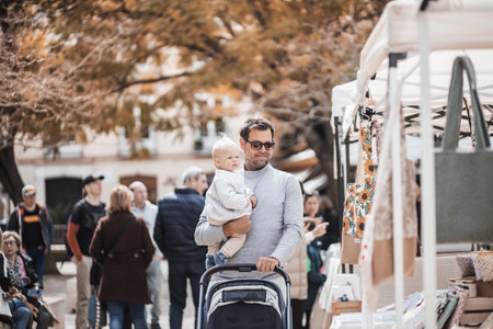Father walking carrying his infant baby boy child and pushing stroller in crowd of people wisiting sunday flea market in Malaga, Spain.の写真素材