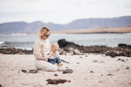 Mother enjoying winter beach vacations playing with his infant baby boy son on wild volcanic sandy beach on Lanzarote island, Canary Islands, Spain. Family travel and vacations concept.の写真素材