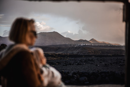 Mother enjoying winter vacations playing with his infant baby boy son on black sandy volcanic beach of Janubio on Lanzarote island, Spain on windy overcast day. Focus on landscape in backgroundの写真素材