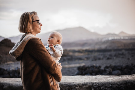 Mother enjoying winter vacations playing with his infant baby boy son on black sandy volcanic beach of Janubio on Lanzarote island, Spain on windy overcast day. Family travel vacations conceptの写真素材